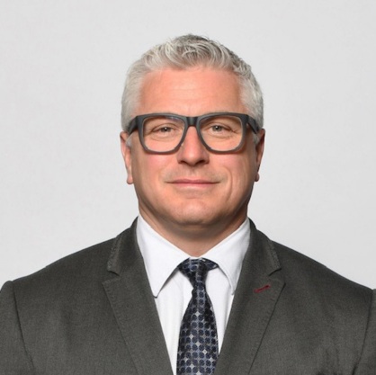 Chris Main smiling with a white button up shirt and gray blazer and gray glasses